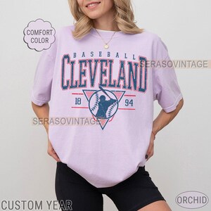 Puede incluir: Una camiseta lavanda con un gr&aacute;fico de b&eacute;isbol vintage. El gr&aacute;fico presenta a un jugador de b&eacute;isbol en un tri&aacute;ngulo con el texto "BASEBALL CLEVELAND 18 94" sobre el jugador.