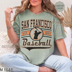 May include: A sage green t-shirt with a vintage baseball graphic. The graphic features a baseball player in a circle with the text "San Francisco Baseball Est. 1883".