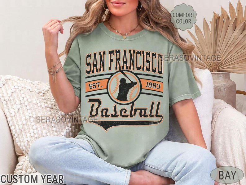 May include: A sage green t-shirt with a vintage baseball graphic. The graphic features a baseball player in a circle with the text "San Francisco Baseball Est. 1883".