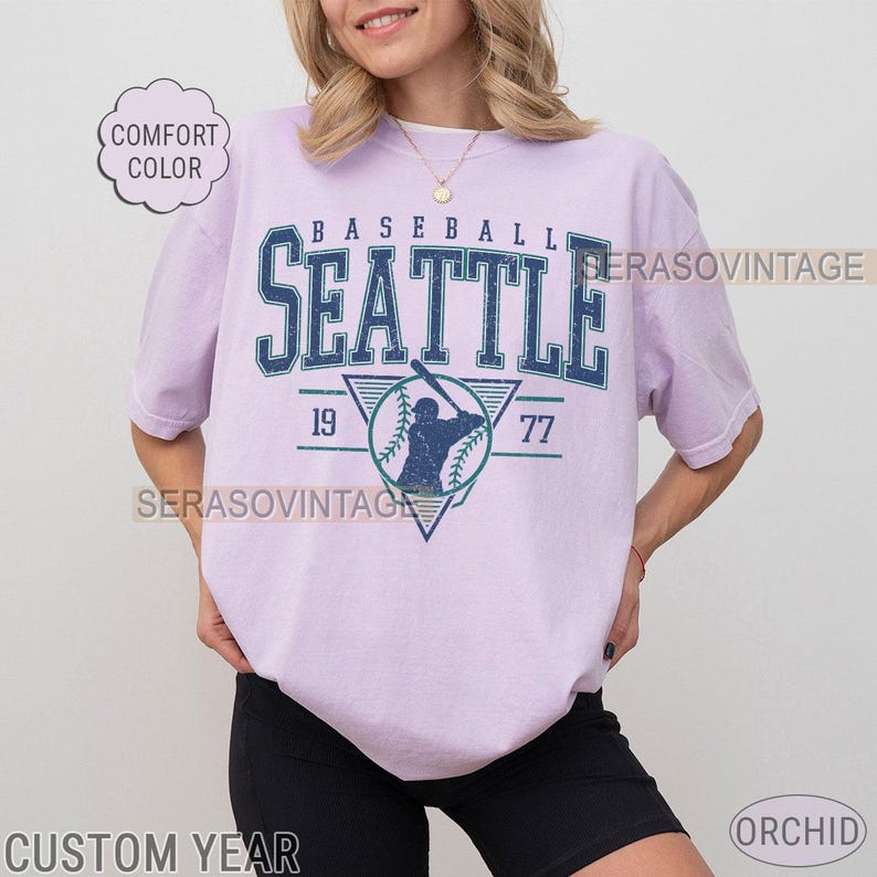 May include: A lavender t-shirt with a vintage baseball graphic. The graphic features a baseball player in silhouette, a baseball, and the text "Baseball Seattle 19 77".