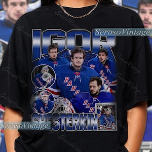 Puede incluir: Camiseta negra con un diseño que presenta a Igor Shesterkin con el uniforme de hockey de los New York Rangers. El diseño incluye el nombre "IGOR CHESTERKIN" en letras azules y blancas, y el logotipo del equipo.