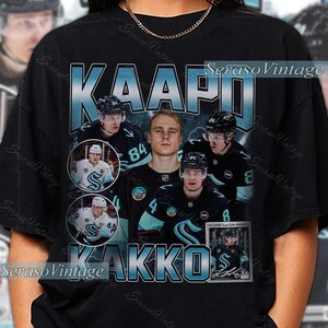 Può includere: T-shirt nera con grafica in stile vintage raffigurante un giocatore di hockey, il nome "KAAPO KAKKO" e il numero 84. Il design in verde acqua e bianco include diverse immagini del giocatore.