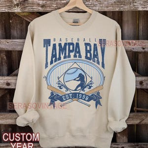 Pode incluir: Camisola de cor bege com um gráfico de beisebol azul e branco. O gráfico apresenta um jogador de beisebol, bastões e o texto "Baseball Tampa Bay Est. 1990".