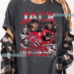 May include: Dark gray t-shirt featuring a graphic of hockey player Jack Hughes. The design includes the name "JACK HUGHES" in red, with images of the player in action. The shirt has a vintage, distressed look.