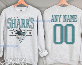 May include: A grey sweatshirt with a San Jose Sharks logo on the front and a blank space on the back for a custom name and number. The logo features a teal shark with a hockey stick and the text "San Jose Sharks" with the years "19" and "91" on either side. The back of the sweatshirt has a teal number "00" and the text "Any Name" above it.