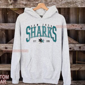 May include: A gray hooded sweatshirt with the words "San Jose Sharks" in teal and white lettering. The logo of the San Jose Sharks hockey team is printed below the text with the words "Est. 1991".