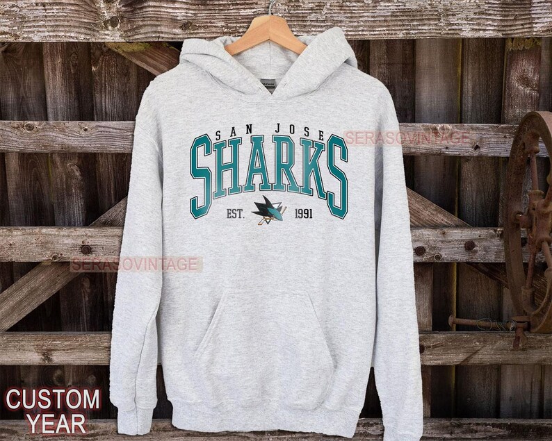 May include: A gray hooded sweatshirt with the words "San Jose Sharks" in teal and white lettering. The logo of the San Jose Sharks hockey team is printed below the text with the words "Est. 1991".