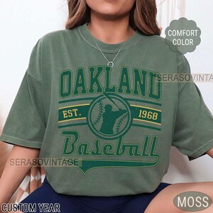 May include: A green t-shirt with a vintage-style graphic of a baseball player and the text "Oakland Baseball Est. 1968".