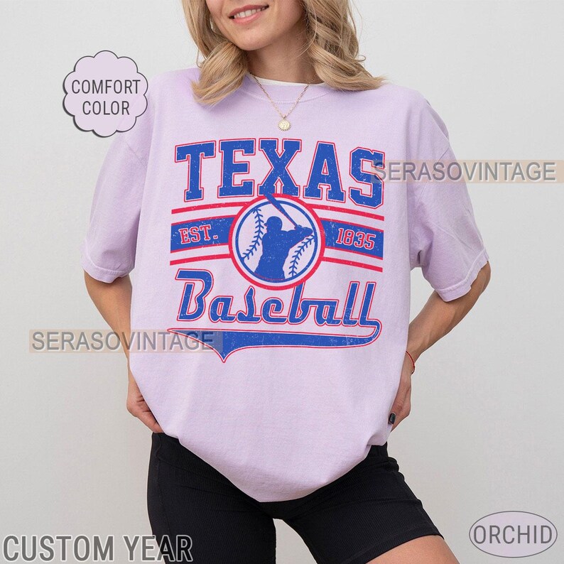 Pode incluir: Uma camiseta lavanda com um gr&aacute;fico de beisebol vintage. O gr&aacute;fico apresenta o texto "Texas Baseball" com os anos "EST. 1835" e uma silhueta de um jogador de beisebol.