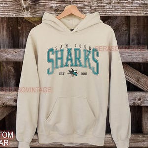 May include: A beige hoodie with a teal blue San Jose Sharks logo and the text "San Jose Sharks Est. 1991".