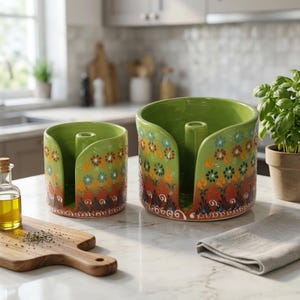 May include: Two green ceramic napkin holders with floral patterns in orange, blue, and yellow. The holders have a curved design with a central support. They sit on a white countertop in a kitchen setting.