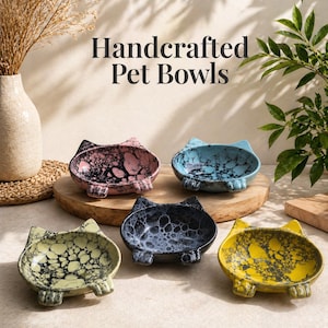 May include: Handcrafted pet bowls in various colors, including pink, blue, black, yellow, and green. Each bowl is cat-shaped with ear-like protrusions and foot-shaped supports. The bowls have a speckled, marbled design.