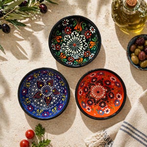 Puede incluir: Tres cuencos de cerámica decorativos con intrincados diseños florales en azul, rojo y verde. Cada cuenco tiene un borde negro y un diseño único. Los cuencos están dispuestos sobre una superficie de color claro, con aceitunas y aceite de oliva cerca.