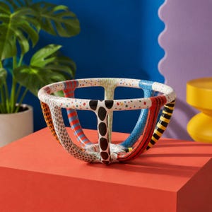 May include: A decorative open-frame bowl featuring a series of curved vertical ribs connected by a circular rim. Each rib displays a distinct pattern, including black spots, red and blue stripes, yellow and black bands, and multicolored dots.