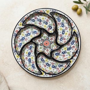 Puede incluir: Una fuente de cerámica blanca con seis compartimentos y una sección central en forma de estrella. La fuente está decorada con coloridos motivos florales en rojo, azul, amarillo y verde, con un borde negro.