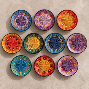 May include: A collection of ten circular ceramic graters featuring raised central grinding surfaces and colorful hand-painted floral patterns in shades of blue, purple, orange, red, yellow, and green, arranged in a grid on a neutral surface.