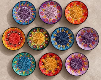May include: A collection of ten circular ceramic graters featuring raised central grinding surfaces and colorful hand-painted floral patterns in shades of blue, purple, orange, red, yellow, and green, arranged in a grid on a neutral surface.