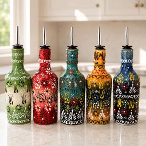 May include: Five glass oil dispensers with silver-colored metal pour spouts. Each bottle features a unique hand-painted floral pattern in vibrant colors including green, red, teal, yellow, and dark blue, with white and black decorative details.