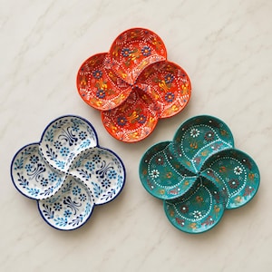 May include: Three four-sectioned ceramic serving dishes arranged in a cluster. One dish is orange with blue and yellow floral patterns, one is white with blue floral motifs, and one is teal with white, red, and yellow floral designs.