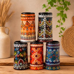 Pode incluir: Cinco copos cilíndricos de cerâmica dispostos numa pilha piramidal. Cada recipiente apresenta padrões intrincados em relevo com cores vibrantes, incluindo laranja, preto, vermelho e azul, decorados com motivos florais e bordas geométricas.