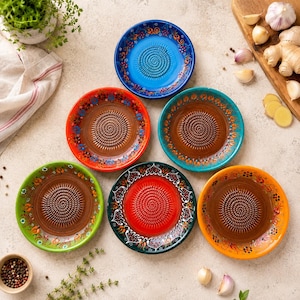 May include: Six circular ceramic graters featuring a central textured grinding surface surrounded by a colorful rim decorated with floral patterns in shades of blue, orange, teal, green, red, and yellow, arranged in a cluster on a surface.