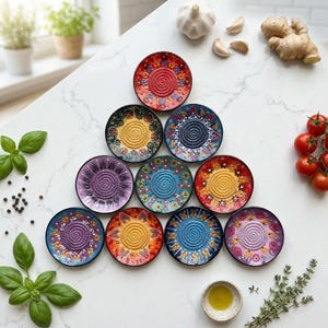 Peut inclure: Dix râpes en céramique colorées avec des motifs floraux et géométriques sont disposées sur un comptoir blanc. Des herbes fraîches, des tomates, de l'ail et du gingembre sont également présents. Chaque assiette a une palette de couleurs unique, dont le rouge, le bleu et le violet.