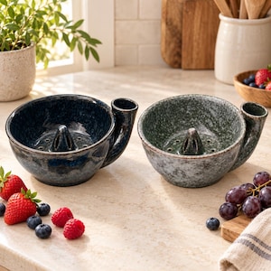 May include: Two ceramic berry colander bowls with a speckled glaze finish, one in deep blue and one in sage green. Each bowl features a central raised cone with drainage holes and a side handle, designed for washing and serving fresh fruit.
