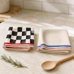 May include: Two square ceramic dishes with folded edges. One dish features a black and white checkerboard pattern with a red striped border. The other dish is solid cream with a blue and red striped border. A wooden spoon and rosemary sprig.