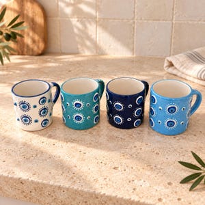 May include: Four ceramic mugs arranged in a row, each featuring a hand-painted evil eye pattern. The mugs are white, teal, dark blue, and light blue, all decorated with concentric circles in blue, white, and black, with a handle on the side.