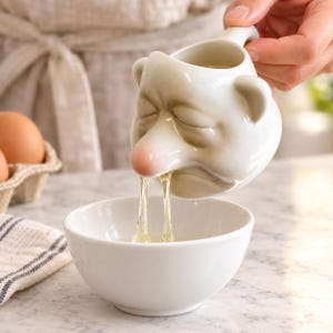 May include: A white ceramic egg separator shaped like a face with closed eyes and a pink nose. The egg white flows from the nostrils of the face into a plain white ceramic bowl. The separator features a small handle on the side for easy pouring.