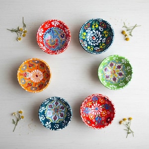 Puede incluir: Colección de siete cuencos pequeños de cerámica pintados a mano. Cada cuenco presenta un diseño floral único y colorido con tonos rojos, verdes, azules y naranjas. Los cuencos están dispuestos en un patrón circular, mostrando sus intrincados diseños y colores vibrantes.