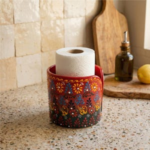 Puede incluir: Un portarrollos de cerámica roja con un diseño floral en amarillo, azul y blanco. Contiene un rollo de papel higiénico blanco. El soporte está sobre una encimera moteada.