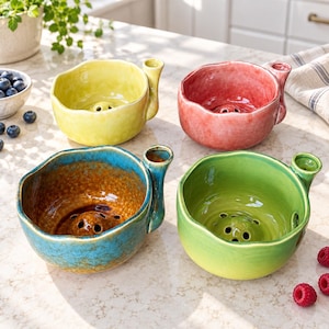 May include: Four ceramic berry bowls in yellow, red, blue, and green. Each bowl features a rounded, organic shape with a small side handle and a raised, perforated center base for drainage. The interior surfaces have a glossy, textured finish.