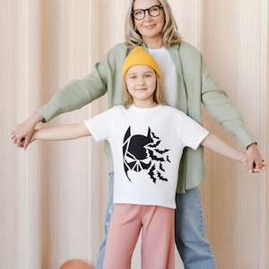 May include: A white t-shirt with a black Batman graphic and bats flying out of the top of the head. A young girl wearing the shirt is holding hands with an adult woman wearing a green shirt.