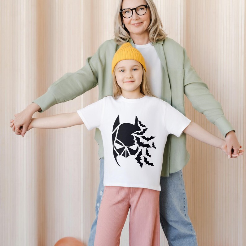 May include: A white t-shirt with a black Batman graphic and bats flying out of the top of the head. A young girl wearing the shirt is holding hands with an adult woman wearing a green shirt.