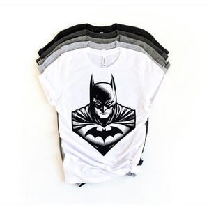 May include: A white t-shirt with a black and white illustration of Batman's face on the front. The shirt is folded and stacked on top of other shirts in gray and black.