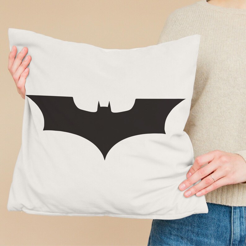 May include: White pillow with a black Batman logo. The logo is a stylized bat silhouette with wings spread wide.