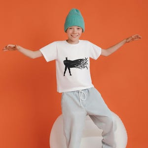 May include: A young person wearing a white t-shirt with a black silhouette of Batman on it. The person is wearing a teal beanie and light grey pants. The person is standing on a white surface with an orange background.