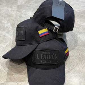 Può includere: Una pila di cappellini da baseball neri con una toppa rettangolare recante la scritta EL PATRON COLOMBIA MEDELLIN in rilievo nero. Ogni cappellino include una piccola toppa ricamata con la bandiera colombiana e un cinturino regolabile.