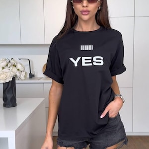 Peut inclure: Un t-shirt noir avec un code-barres blanc et le mot "YES" imprimé sur le devant. Le mannequin porte le t-shirt avec un short en jean déchiré.