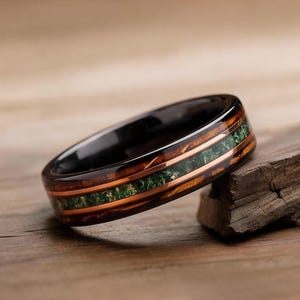 May include: A black ring with a wood grain and green stone inlay. The ring has a polished finish and is set on a piece of wood. The ring is a unique piece of jewellery.