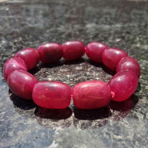 May include: A circular bracelet composed of smooth, polished, cylindrical beads in a deep, vibrant magenta-red color. The beads are strung together in a continuous loop, showcasing a consistent glossy finish and uniform shape throughout the piece.