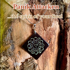 Könnte beinhalten: Eine silberne Halskette mit einem dunklen quadratischen Anhänger mit einem weißen floralen Design und dem Text "Panik Attacken...the spirit of your Soul". Der Name des Künstlers, Ina Adreju, ist ebenfalls sichtbar.