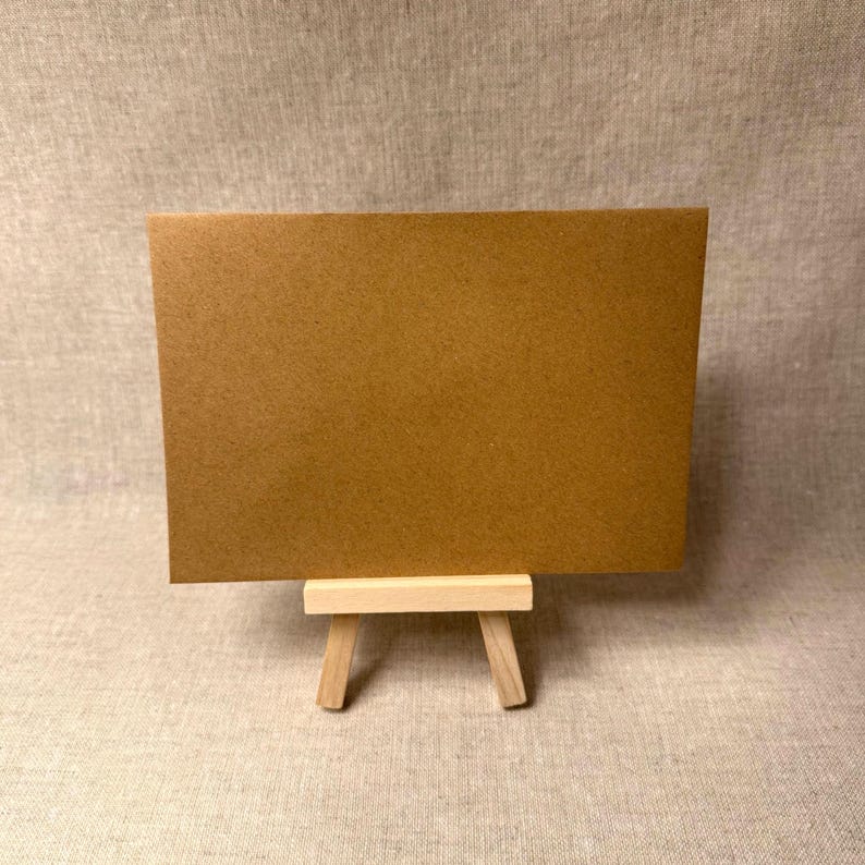 May include: A brown rectangular card displayed on a small wooden easel. The card has a smooth surface and is a warm, earthy tone. The easel is made of light-colored wood and has a simple, minimalist design. The background is a neutral fabric.