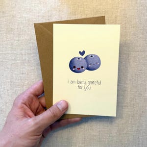 May include: A greeting card with a light yellow background and a brown envelope. The card features two cartoon blueberries with smiling faces and a small blue heart. The text reads, "I am berry grateful for you."
