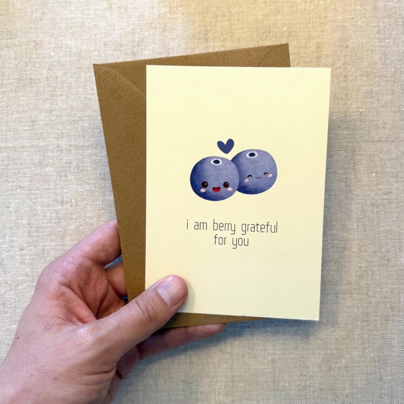 May include: A greeting card with a light yellow background and a brown envelope. The card features two cartoon blueberries with smiling faces and a small blue heart. The text reads, "I am berry grateful for you."