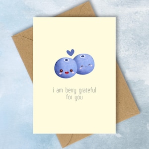 May include: A greeting card with a light yellow background and a brown envelope. The card features two cartoon blueberries with smiling faces and a small blue heart. The text reads, "i am berry grateful for you."