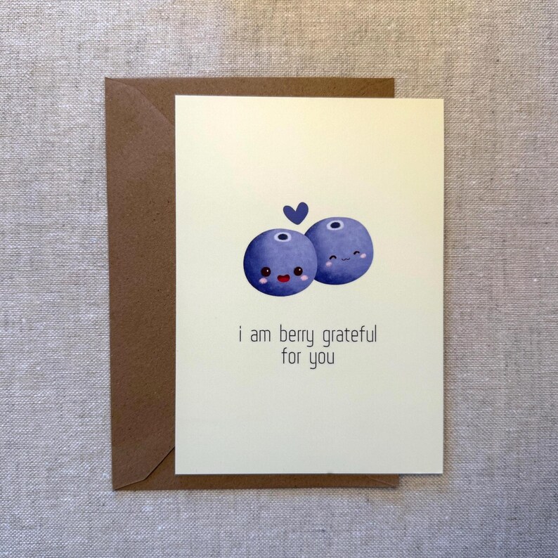 May include: A greeting card with a light cream background and a brown envelope. The card features two cartoon blueberries with smiling faces and a small blue heart. The text reads, "i am berry grateful for you."