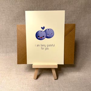 May include: A greeting card with a watercolor illustration of two smiling blueberries and a small blue heart. The text "i am berry grateful for you" is printed below the blueberries. The card is displayed on a small wooden easel, with a brown envelope behind it.