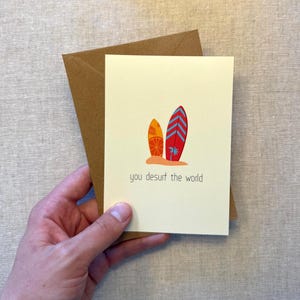 Könnte beinhalten: Grußkarte mit zwei Surfbrettern am Strand, eines orange und eines rot, mit dem Text "you desurf the world". Die Karte wird mit einem braunen Umschlag gehalten.